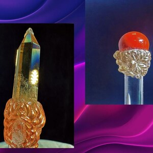 Puede incluir: Dos objetos decorativos de cristal. Uno es un cristal alto y facetado con un tono dorado, colocado sobre una base ornamentada y tallada. El otro es una varilla transparente coronada por una esfera roja, sujeta por un dise&ntilde;o dorado en forma de hoja.