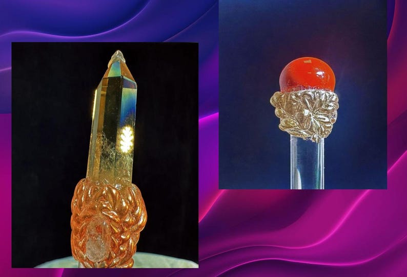 Puede incluir: Dos objetos decorativos de cristal. Uno es un cristal alto y facetado con un tono dorado, colocado sobre una base ornamentada y tallada. El otro es una varilla transparente coronada por una esfera roja, sujeta por un dise&ntilde;o dorado en forma de hoja.