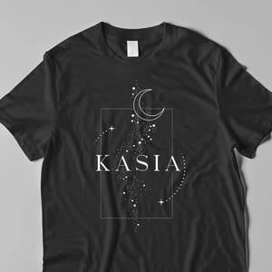 Puede incluir: Camiseta negra con un diseño celestial blanco. El diseño incluye una luna creciente, estrellas y el nombre "KASIA" en un marco rectangular. La camiseta está hecha de un material suave y es adecuada para uso casual.