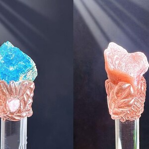Puede incluir: Dos esculturas decorativas de cristal. Una presenta un cristal azul vibrante sobre una base detallada de color rosa. La otra muestra un cristal color melocot&oacute;n sobre una base similar. Ambas est&aacute;n montadas sobre soportes cil&iacute;ndricos transparentes.
