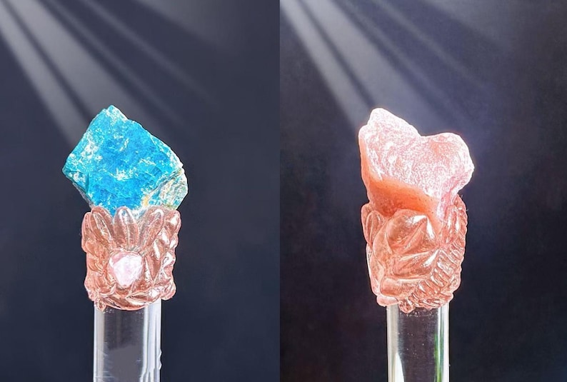 Puede incluir: Dos esculturas decorativas de cristal. Una presenta un cristal azul vibrante sobre una base detallada de color rosa. La otra muestra un cristal color melocot&oacute;n sobre una base similar. Ambas est&aacute;n montadas sobre soportes cil&iacute;ndricos transparentes.