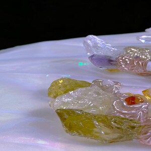 Puede incluir: Primer plano de varitas de cristal con varias piedras preciosas de colores. Las varitas presentan cristales transparentes, amarillos, morados y rosas. Las varitas est&aacute;n sobre una superficie blanca e iridiscente con un fondo oscuro.