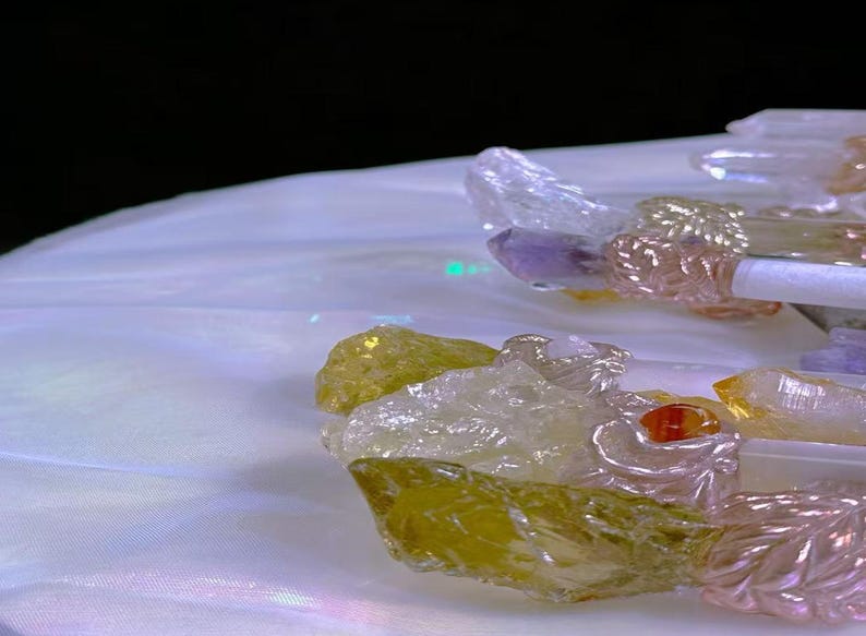 Puede incluir: Primer plano de varitas de cristal con varias piedras preciosas de colores. Las varitas presentan cristales transparentes, amarillos, morados y rosas. Las varitas est&aacute;n sobre una superficie blanca e iridiscente con un fondo oscuro.