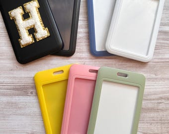 May include: A collection of rectangular vertical badge holders in black, blue, white, yellow, pink, and sage green. One black holder features a white letter H patch with a gold glitter border. Each holder has a clear window and a top slot.