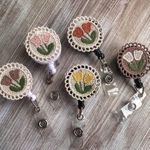 May include: A collection of five floral embroidered badge reels. Each reel features a circular design with a crocheted border and a floral motif in various colors, including pink, orange, yellow, and brown. The reels have a retractable cord and a clear plastic clip.