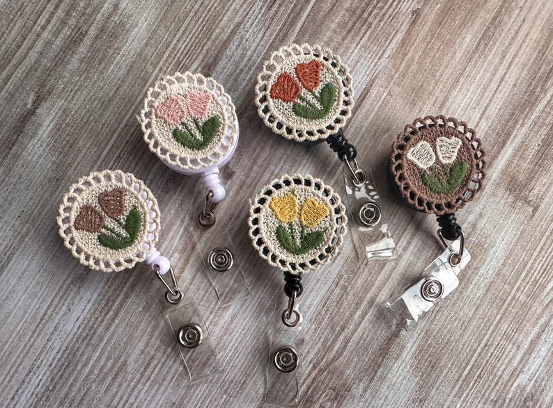 May include: A collection of five floral embroidered badge reels. Each reel features a circular design with a crocheted border and a floral motif in various colors, including pink, orange, yellow, and brown. The reels have a retractable cord and a clear plastic clip.