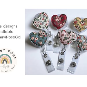 May include: Heart-shaped badge reels with floral fabric designs in various colors, including red, pink, and blue. The reels are attached to clear plastic retractable holders. Text on the image reads "More designs available at HenryRoseCo!"