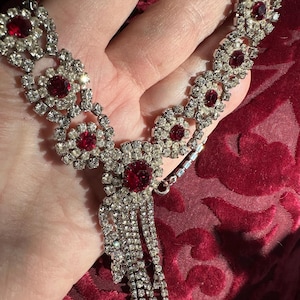 May include: A silver-toned necklace with red and clear gemstones. The necklace features a series of floral-shaped clusters with a large red gemstone in the center of each. A cascade of clear gemstones hangs from the center.