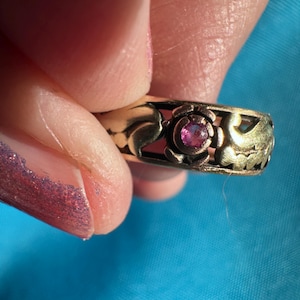Pode incluir: Um anel de metal em tom dourado com uma banda vazada e um motivo floral repetitivo. O centro do design da flor é cravejado com uma pequena pedra preciosa redonda de cor rosa vibrante. A banda de metal tem um acabamento texturizado.