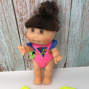 May include: A Cabbage Patch Kid doll wearing a pink swimsuit with a blue and green trim. The swimsuit has a rainbow fish design and the text "CPR" on the front. The doll has brown hair and brown eyes. The doll is also wearing a yellow life vest with pink trim and a blue strap.