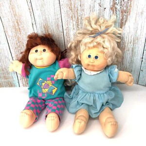 Puede incluir: Dos muñecas Cabbage Patch Kids, una con cabello castaño que lleva una camisa turquesa con un patrón geométrico rosa y azul y pantalones a cuadros rosa y turquesa, y la otra con cabello rubio que lleva un vestido azul claro con un borde de encaje blanco.