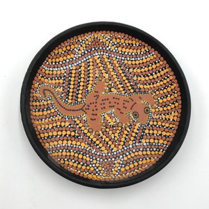 May include: A black ceramic plate with a brown and white dot pattern and a brown gecko design. The gecko is in the center of the plate and is facing to the right.
