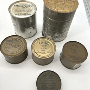 May include: An assortment of vintage metal food cans, including baking powder, crackers, cocoa beverage powder, and peanut butter. The cans are various sizes and shapes, with labels and text visible. The labels include brand names and product information.