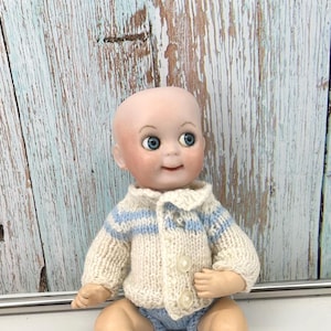 May include: A vintage baby doll with a bald head, blue eyes, and rosy cheeks. The doll wears a cream-colored knitted sweater with blue stripes and matching blue shorts. The doll is sitting down.