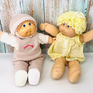 Może przedstawiać: Dwie lalki Cabbage Patch Kids, jedna ubrana w różowo-biały dzianinowy strój i białą czapkę, druga w żółtą sukienkę z białą kokardką. Obie lalki mają blond włosy i niebieskie oczy.