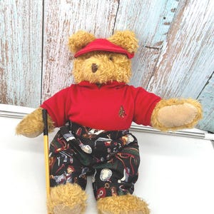 May include: A plush teddy bear dressed in a red golf shirt and cap, with patterned golf pants. The bear is holding a miniature golf club. The bear's fur is light brown, and the outfit has a golf theme.