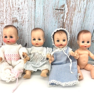May include: Four vintage baby dolls with painted facial features and rooted hair. The dolls are dressed in various outfits, including a white knit sweater, a blue and white gingham dress, and a striped dress. One doll is undressed. The dolls are sitting on a white surface.