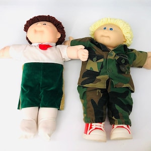 Könnte beinhalten: Zwei Cabbage Patch Kids Puppen, eine mit braunen Haaren, die ein weißes Hemd mit einer roten Schleife und eine grüne Hose trägt, und die andere mit blonden Haaren, die ein Tarnhemd und eine Tarnhose mit roten Schuhen trägt.