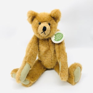 May include: A brown teddy bear with a green tag that reads "The Green Mountain Bears" and "Hand Made" is sitting on a white surface.