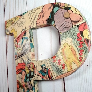 Puede incluir: Una letra P de madera cubierta con un collage de p&aacute;ginas de c&oacute;mics que muestran a un superh&eacute;roe con un martillo. El texto "THE PAIN IS STILL GREAT" es visible en la letra.
