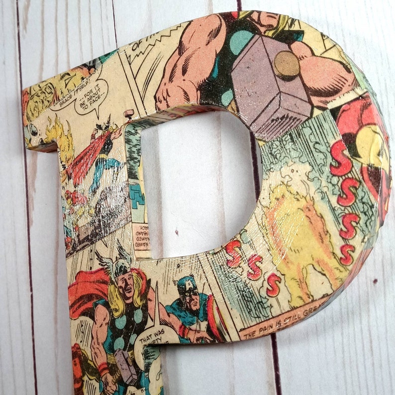 Puede incluir: Una letra P de madera cubierta con un collage de p&aacute;ginas de c&oacute;mics que muestran a un superh&eacute;roe con un martillo. El texto "THE PAIN IS STILL GREAT" es visible en la letra.