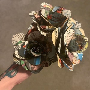 K&ouml;nnte beinhalten: Ein Strau&szlig; aus drei Papierrosen, die aus Comic-Seiten hergestellt wurden. Die Rosen sind dunkelbraun, schwarz und wei&szlig;. Die Comic-Seiten zeigen Bilder von Superhelden und Text.