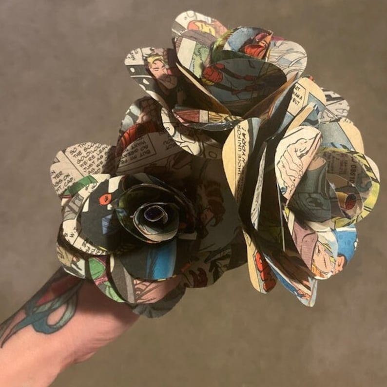 K&ouml;nnte beinhalten: Ein Strau&szlig; aus drei Papierrosen, die aus Comic-Seiten hergestellt wurden. Die Rosen sind dunkelbraun, schwarz und wei&szlig;. Die Comic-Seiten zeigen Bilder von Superhelden und Text.