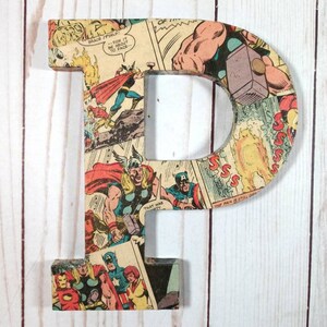 Puede incluir: Una gran letra P de madera cubierta de p&aacute;ginas de c&oacute;mics con superh&eacute;roes como Thor y Capit&aacute;n Am&eacute;rica. La letra est&aacute; pintada de color marr&oacute;n claro y tiene un aspecto desgastado.