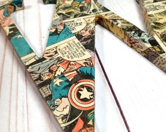 May include: A large, wooden letter V covered in vintage comic book pages featuring superheroes. The letter is painted in a light brown color and has a distressed finish. The comic book pages are a mix of colors, including red, blue, yellow, and green. The letter is standing upright on a white wooden surface.