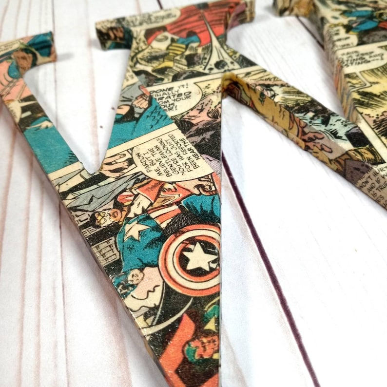 Puede incluir: Una gran letra V de madera cubierta de p&aacute;ginas de c&oacute;mics vintage con superh&eacute;roes. La letra est&aacute; pintada de color marr&oacute;n claro y tiene un acabado desgastado. Las p&aacute;ginas de c&oacute;mics son una mezcla de colores, incluyendo rojo, azul, amarillo y verde. La letra est&aacute; de pie sobre una superficie de madera blanca.