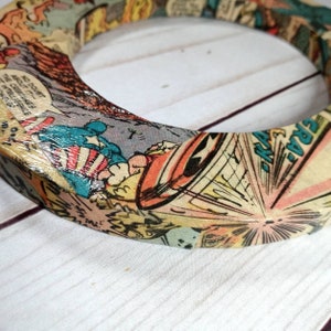 Puede incluir: Un anillo de madera con un dise&ntilde;o de portada de c&oacute;mic. El anillo est&aacute; decorado con una imagen colorida de un superh&eacute;roe volando por el aire. El texto "NO DOUBT ABOUT IT!" es visible en el anillo.