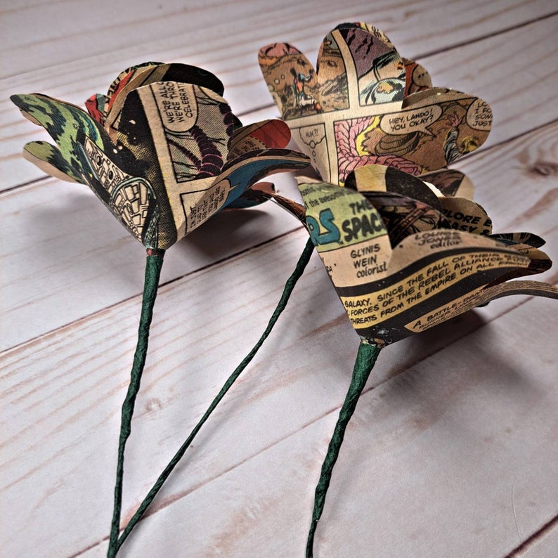 K&ouml;nnte beinhalten: Drei Papierrosen aus alten Comic-Seiten. Die Rosen sind hellbraun mit schwarzem und wei&szlig;em Text und Bildern. Die Stiele sind in gr&uuml;nes Papier gewickelt.