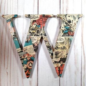 Puede incluir: Dos letras de madera, W y V, cubiertas con p&aacute;ginas de c&oacute;mics. Las letras est&aacute;n pintadas de color marr&oacute;n claro y tienen un aspecto desgastado. Las p&aacute;ginas de c&oacute;mics presentan superh&eacute;roes y escenas de acci&oacute;n.