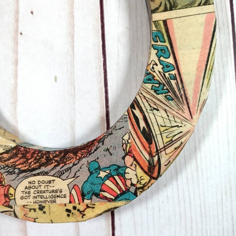 Puede incluir: Un posavasos circular de madera con un dise&ntilde;o de c&oacute;mic que presenta a Capit&aacute;n Am&eacute;rica. El posavasos tiene un esquema de color rojo, blanco y azul y presenta el texto "NO DOUBT ABOUT IT - THE CREATURE'S GOT INTELLIGENCE - HOWEVER".