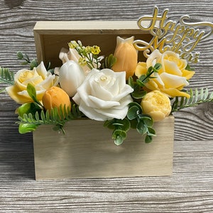 May include: A wooden box filled with a bouquet of artificial flowers. The flowers are white and yellow roses, tulips, and other blooms. The box is decorated with a sign that says "Happy Mother's".