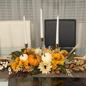 May include: A fall-themed centerpiece featuring three white candles in gold holders, surrounded by pumpkins, yellow flowers, and decorative foliage. The arrangement sits on a dark brown runner, creating a festive table decoration.