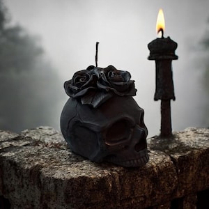 May include: A black skull-shaped candle with a cluster of black roses on top, resting on a stone surface. A lit candle with a flame is to the right. The scene has a gothic or Halloween theme.