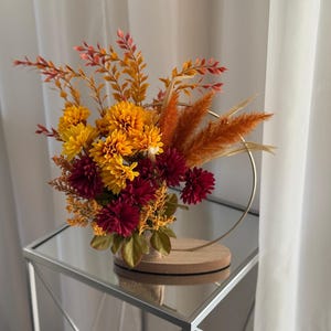 May include: A floral display with yellow, orange, and burgundy blooms, complemented by dried grasses and foliage. The flowers are arranged in a gold metal ring, placed on a wooden base. The arrangement sits on a mirrored surface.