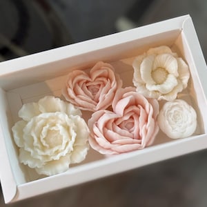 May include: A white box contains six floral-shaped soaps. The soaps are in shades of white and pink, with detailed petal designs. The arrangement includes roses and heart-shaped flowers, creating a delicate and elegant aesthetic.