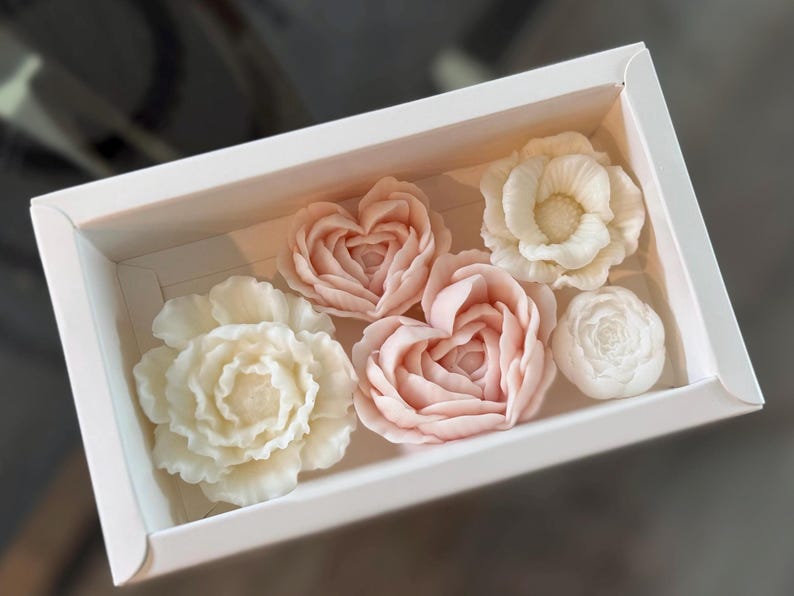 May include: A white box contains six floral-shaped soaps. The soaps are in shades of white and pink, with detailed petal designs. The arrangement includes roses and heart-shaped flowers, creating a delicate and elegant aesthetic.
