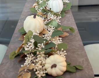 May include: A table runner with cream pumpkins, green leaves, and white berries. The arrangement is on a grey surface, creating a seasonal centrepiece. The pumpkins have brown stems, perfect for autumn decor.