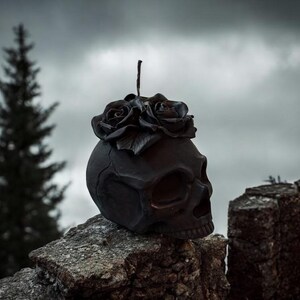 May include: A dark, gothic-themed skull sculpture with a floral crown of roses. The skull is black and rests on a stone surface, set against a cloudy sky. The roses are also black, adding to the somber aesthetic.
