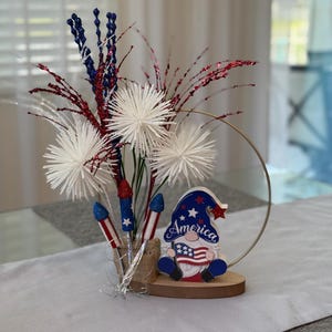 May include: A patriotic centerpiece featuring a wooden gnome holding a flag that says "America" and a vase filled with white and red sparkly firework-like stems.