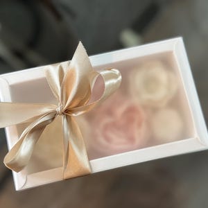 May include: A white rectangular gift box with a clear lid, tied with a gold satin ribbon bow. Inside the box are several light pink and white floral-shaped items, visible through the clear lid.