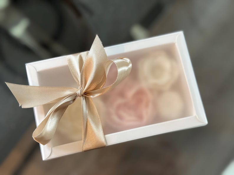 May include: A white rectangular gift box with a clear lid, tied with a gold satin ribbon bow. Inside the box are several light pink and white floral-shaped items, visible through the clear lid.