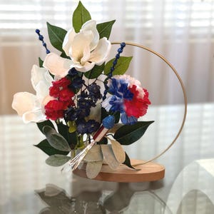 May include: A white, red, and blue flower arrangement in a gold metal hoop stand. The flowers are arranged on a wooden base.
