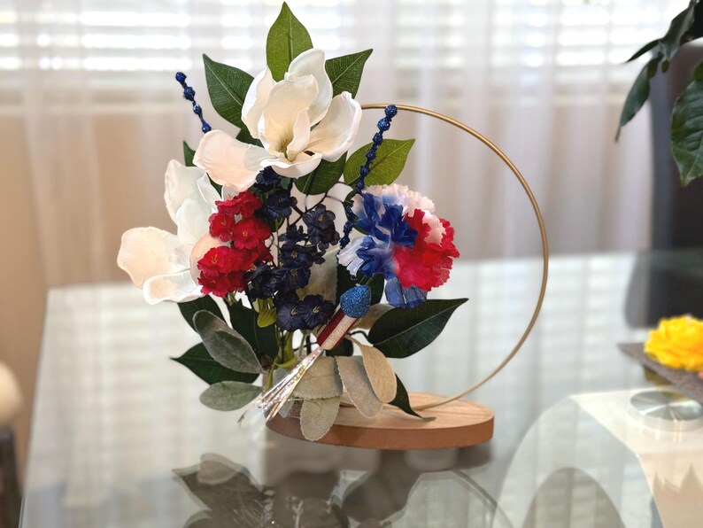 May include: A white, red, and blue flower arrangement in a gold metal hoop stand. The flowers are arranged on a wooden base.