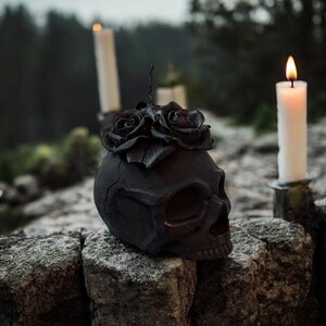 May include: A black skull sculpture adorned with black roses, resting on a stone wall. Lit white candles in holders flank the skull, with a blurred forest background. The scene evokes a gothic or Halloween theme.