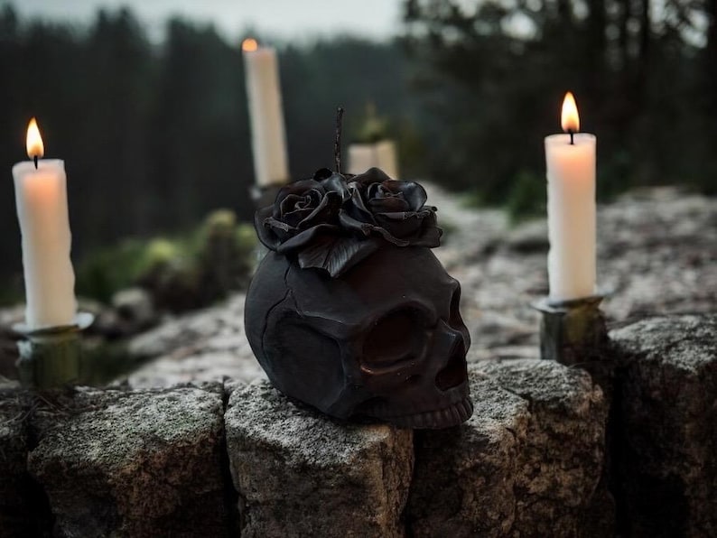 May include: A black skull sculpture adorned with black roses, resting on a stone wall. Lit white candles in holders flank the skull, with a blurred forest background. The scene evokes a gothic or Halloween theme.