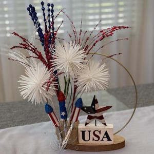 May include: A patriotic centerpiece featuring white, red, and blue fireworks, a star with the American flag design, and a wooden block with the word "USA" on it. The centerpiece is surrounded by a gold metal ring.